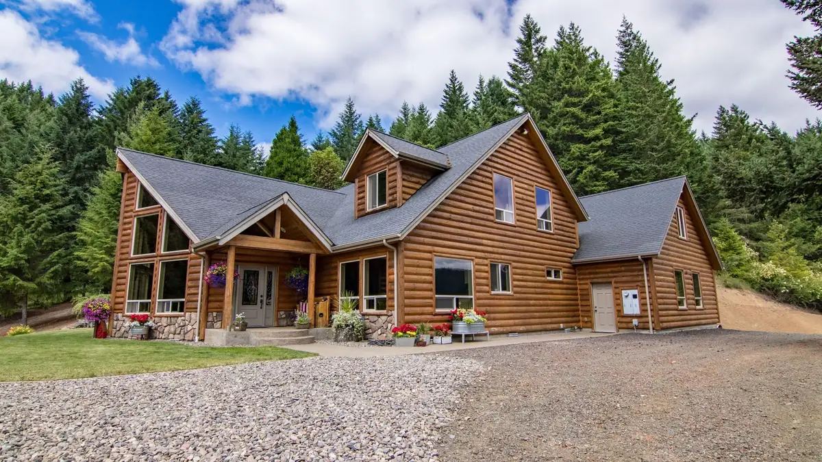 A log cabin with a wide front porch in a clearing, surrounded by tall green trees under a blue sky.